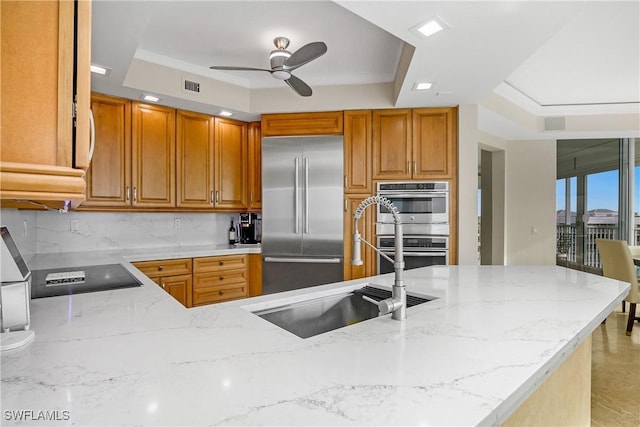 kitchen with appliances with stainless steel finishes, ceiling fan, kitchen peninsula, a raised ceiling, and light stone counters