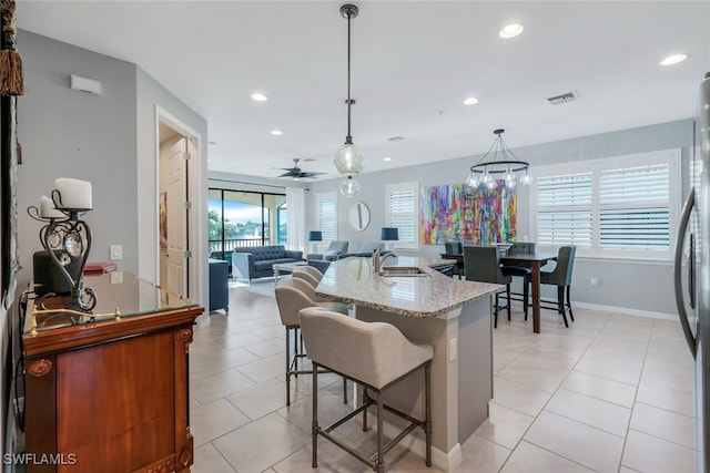 kitchen featuring a kitchen bar, ceiling fan with notable chandelier, light stone countertops, a center island with sink, and pendant lighting