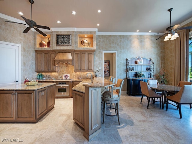 kitchen with premium range hood, an island with sink, appliances with stainless steel finishes, light stone countertops, and crown molding