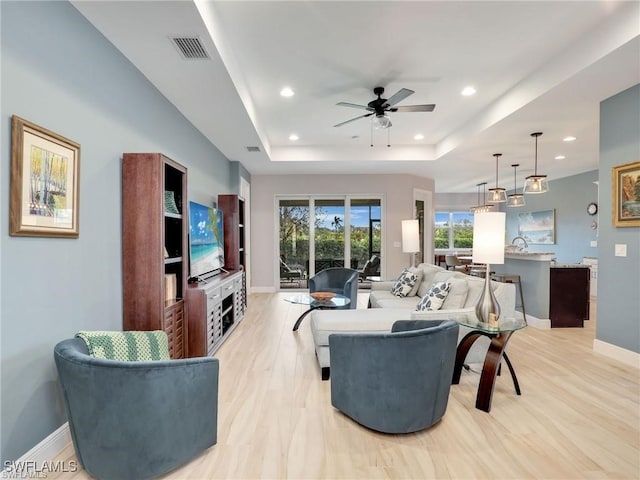 living room with a raised ceiling, ceiling fan, and light wood-type flooring