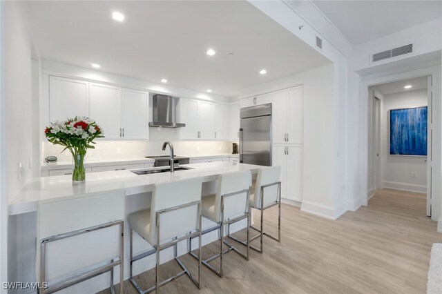 kitchen with a kitchen bar, built in fridge, wall chimney exhaust hood, white cabinets, and sink