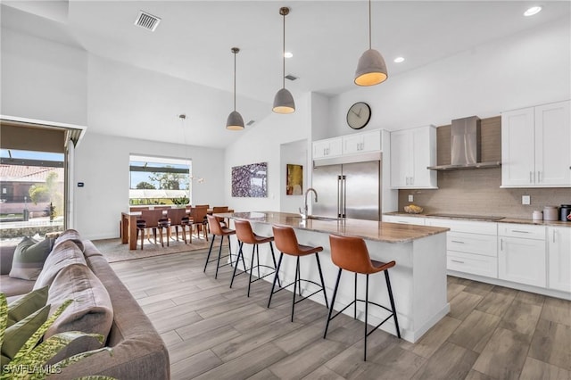 kitchen featuring white cabinets, stainless steel built in fridge, hanging light fixtures, and wall chimney range hood