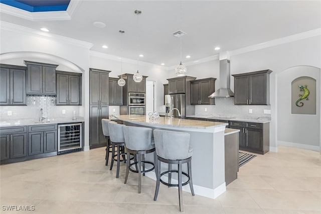 kitchen with wall chimney exhaust hood, beverage cooler, hanging light fixtures, a kitchen island with sink, and appliances with stainless steel finishes