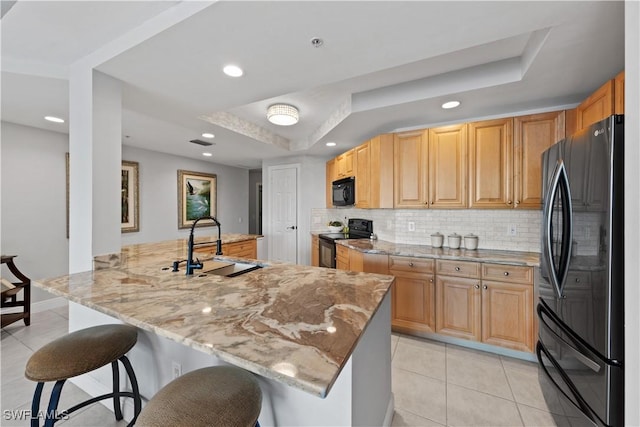 kitchen with kitchen peninsula, light stone countertops, black appliances, a tray ceiling, and sink