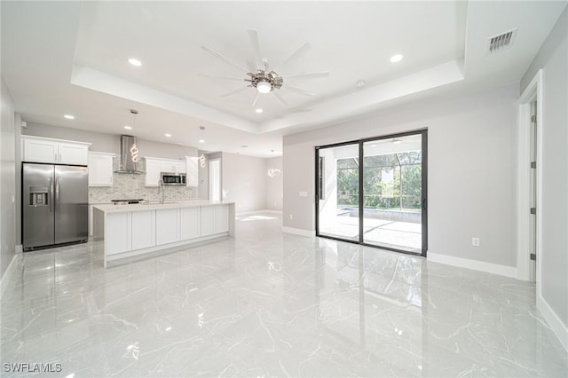 kitchen with appliances with stainless steel finishes, an island with sink, white cabinets, a raised ceiling, and wall chimney exhaust hood