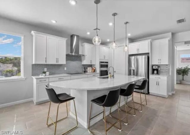 kitchen featuring stainless steel appliances, wall chimney range hood, a kitchen island with sink, a breakfast bar area, and white cabinetry