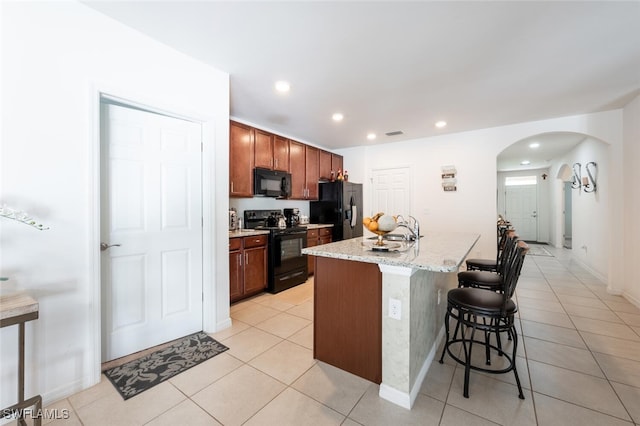 kitchen featuring light stone countertops, a center island with sink, black appliances, light tile patterned flooring, and a kitchen breakfast bar