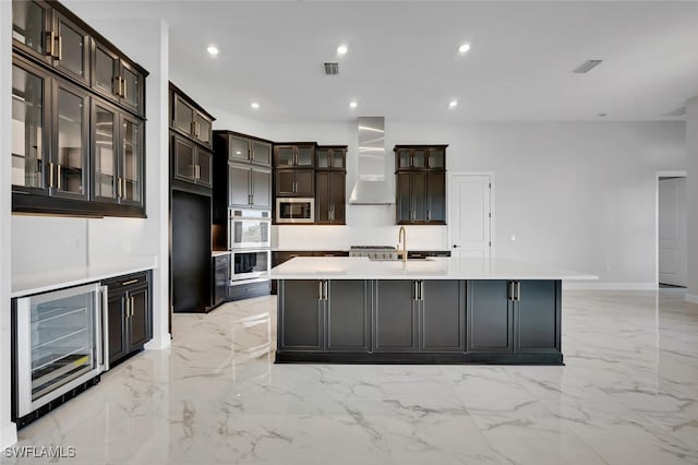 kitchen with beverage cooler, stainless steel microwave, light countertops, wall chimney exhaust hood, and an island with sink