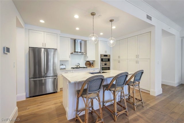 kitchen with light stone countertops, wall chimney exhaust hood, stainless steel appliances, a center island with sink, and white cabinetry