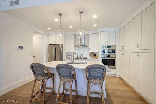 kitchen featuring appliances with stainless steel finishes, white cabinetry, wall chimney range hood, and a kitchen island with sink