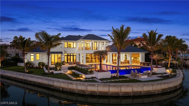 back house at dusk featuring a balcony, a swimming pool with hot tub, a yard, a water view, and a patio area