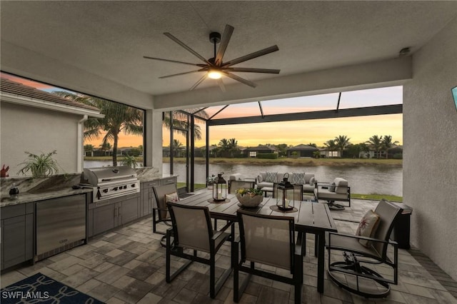 patio terrace at dusk featuring glass enclosure, ceiling fan, exterior kitchen, a grill, and a water view