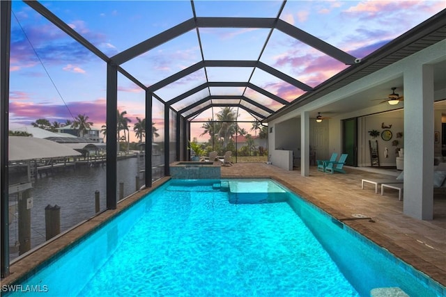 pool at dusk featuring ceiling fan, glass enclosure, a water view, and a patio