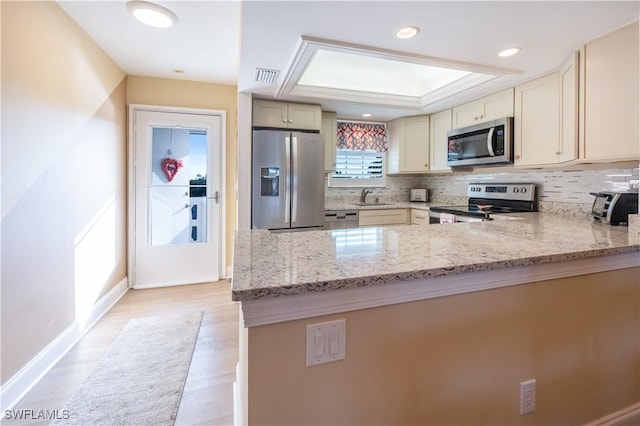 kitchen with cream cabinetry, stainless steel appliances, a raised ceiling, light stone countertops, and sink