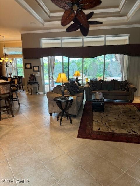 living room with ceiling fan with notable chandelier, a raised ceiling, ornamental molding, and light tile patterned floors