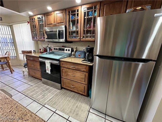 kitchen with backsplash, brown cabinetry, glass insert cabinets, light tile patterned flooring, and stainless steel appliances