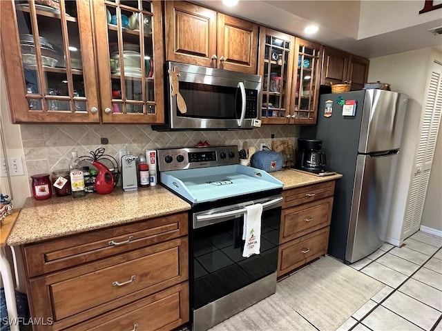 kitchen featuring tasteful backsplash, glass insert cabinets, light tile patterned floors, light stone counters, and appliances with stainless steel finishes