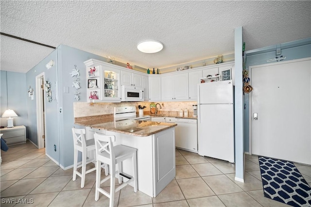 kitchen with light tile patterned flooring, a peninsula, white appliances, white cabinets, and glass insert cabinets