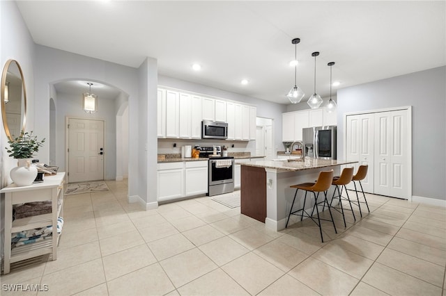 kitchen featuring an island with sink, stainless steel appliances, pendant lighting, white cabinets, and light stone counters