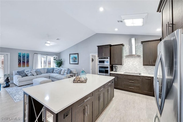 kitchen with wall chimney range hood, appliances with stainless steel finishes, a kitchen island, decorative backsplash, and vaulted ceiling