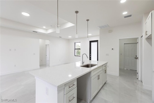 kitchen featuring sink, white cabinetry, light stone counters, hanging light fixtures, and an island with sink