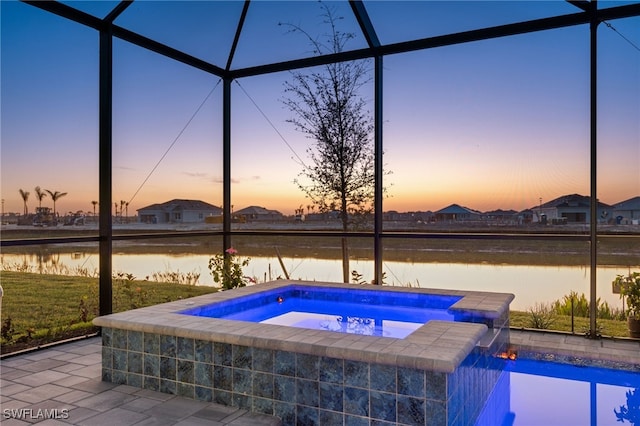 pool at dusk featuring an in ground hot tub, a water view, glass enclosure, and a patio area