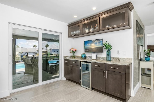 bar with wine cooler, dark brown cabinets, and light stone countertops