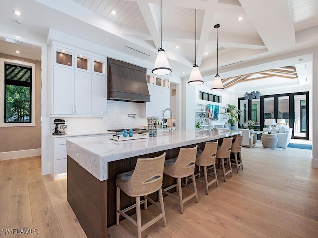 kitchen with a large island with sink, premium range hood, pendant lighting, beamed ceiling, and white cabinets