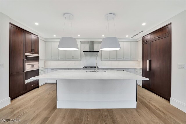 kitchen featuring dark brown cabinetry, wall chimney exhaust hood, light wood-type flooring, pendant lighting, and a kitchen island with sink