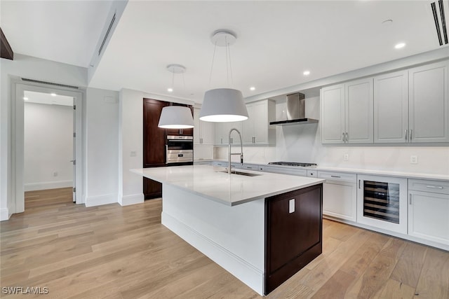 kitchen with wall chimney exhaust hood, sink, hanging light fixtures, an island with sink, and beverage cooler