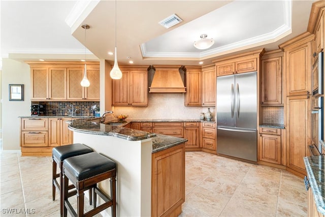 kitchen featuring built in refrigerator, dark stone counters, ornamental molding, a tray ceiling, and custom range hood