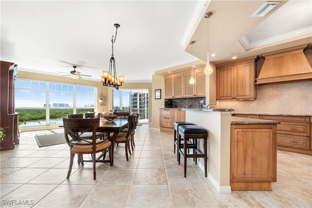 kitchen with ceiling fan with notable chandelier, custom exhaust hood, kitchen peninsula, crown molding, and light stone counters