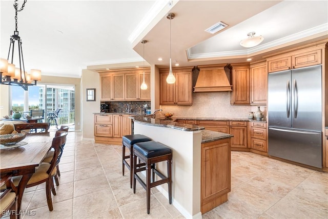 kitchen with hanging light fixtures, stainless steel built in fridge, dark stone countertops, and custom range hood