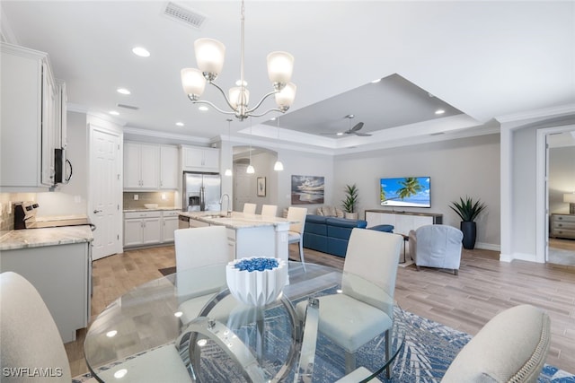 dining room with sink, light hardwood / wood-style floors, ceiling fan with notable chandelier, a tray ceiling, and crown molding