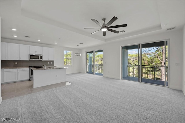 kitchen with a raised ceiling, stainless steel appliances, an island with sink, hanging light fixtures, and white cabinetry