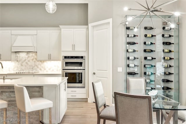 kitchen featuring double oven, white cabinetry, backsplash, light hardwood / wood-style floors, and custom range hood