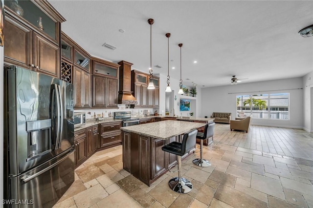 kitchen with stainless steel appliances, custom range hood, decorative light fixtures, a kitchen breakfast bar, and light stone countertops