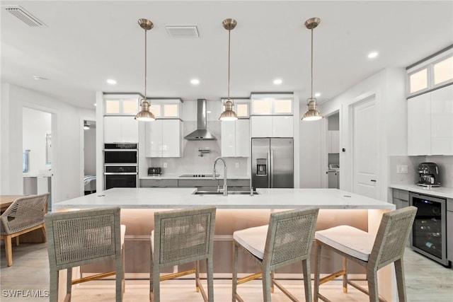 kitchen featuring stainless steel fridge, wine cooler, white cabinetry, wall chimney range hood, and an island with sink