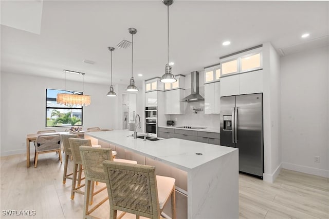 kitchen with white cabinets, decorative light fixtures, an island with sink, stainless steel appliances, and wall chimney exhaust hood