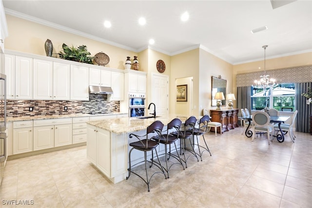 kitchen with light stone counters, decorative light fixtures, ornamental molding, a kitchen breakfast bar, and a kitchen island with sink