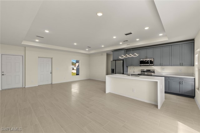 kitchen featuring a sink, a kitchen island with sink, light countertops, appliances with stainless steel finishes, and a raised ceiling
