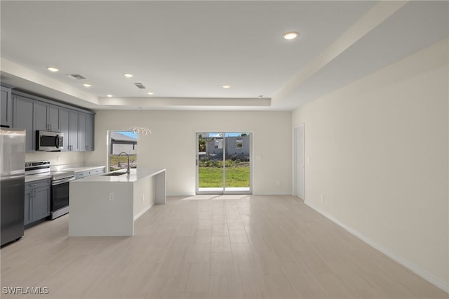 kitchen with sink, a tray ceiling, a kitchen island with sink, appliances with stainless steel finishes, and gray cabinetry