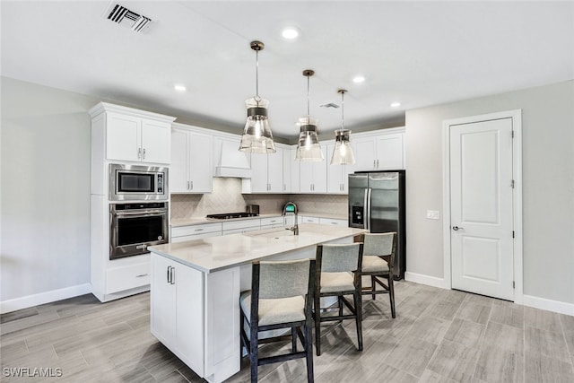 kitchen with appliances with stainless steel finishes, white cabinetry, hanging light fixtures, and an island with sink