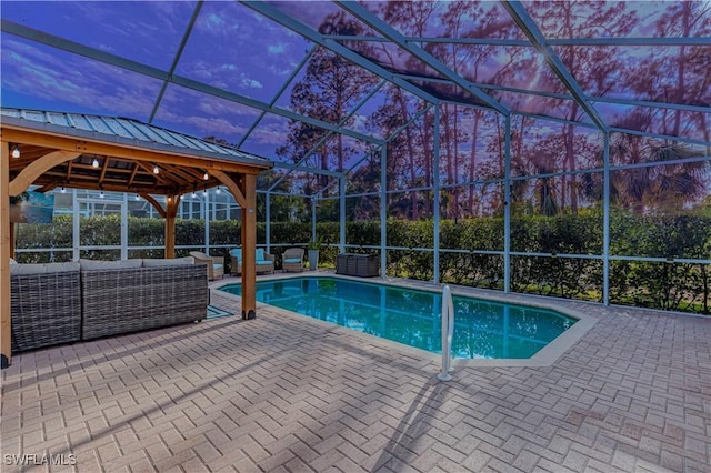 pool at dusk featuring glass enclosure, a patio area, a gazebo, and an outdoor hangout area