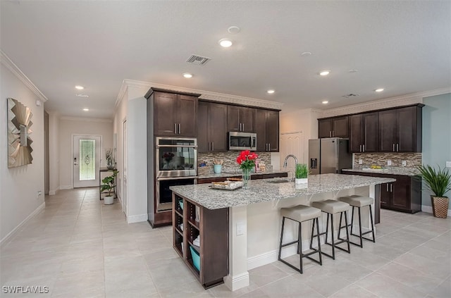 kitchen with dark brown cabinetry, appliances with stainless steel finishes, ornamental molding, and a center island with sink