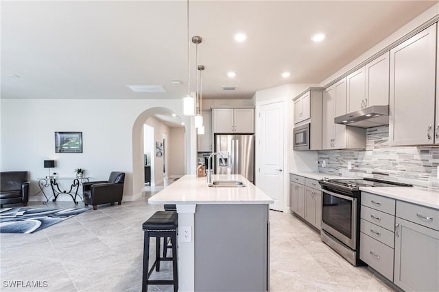 kitchen with sink, gray cabinetry, stainless steel appliances, an island with sink, and decorative light fixtures