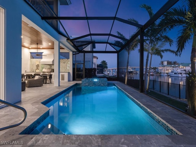 pool at night featuring an in ground hot tub, a lanai, a water view, a patio area, and an outdoor hangout area