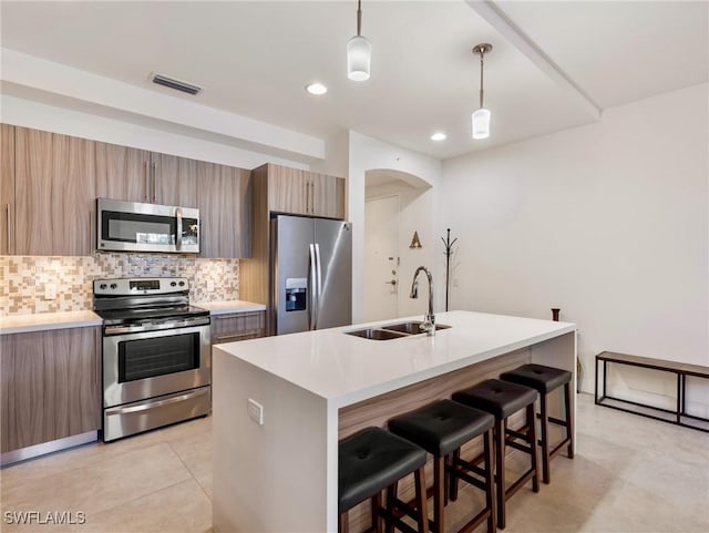 kitchen featuring appliances with stainless steel finishes, backsplash, a kitchen island with sink, decorative light fixtures, and sink