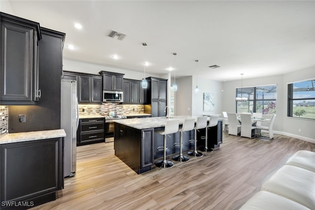 kitchen with an island with sink, stainless steel appliances, tasteful backsplash, decorative light fixtures, and light stone countertops