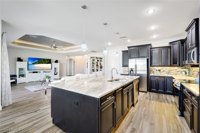 kitchen with pendant lighting, stainless steel appliances, an island with sink, sink, and a tray ceiling